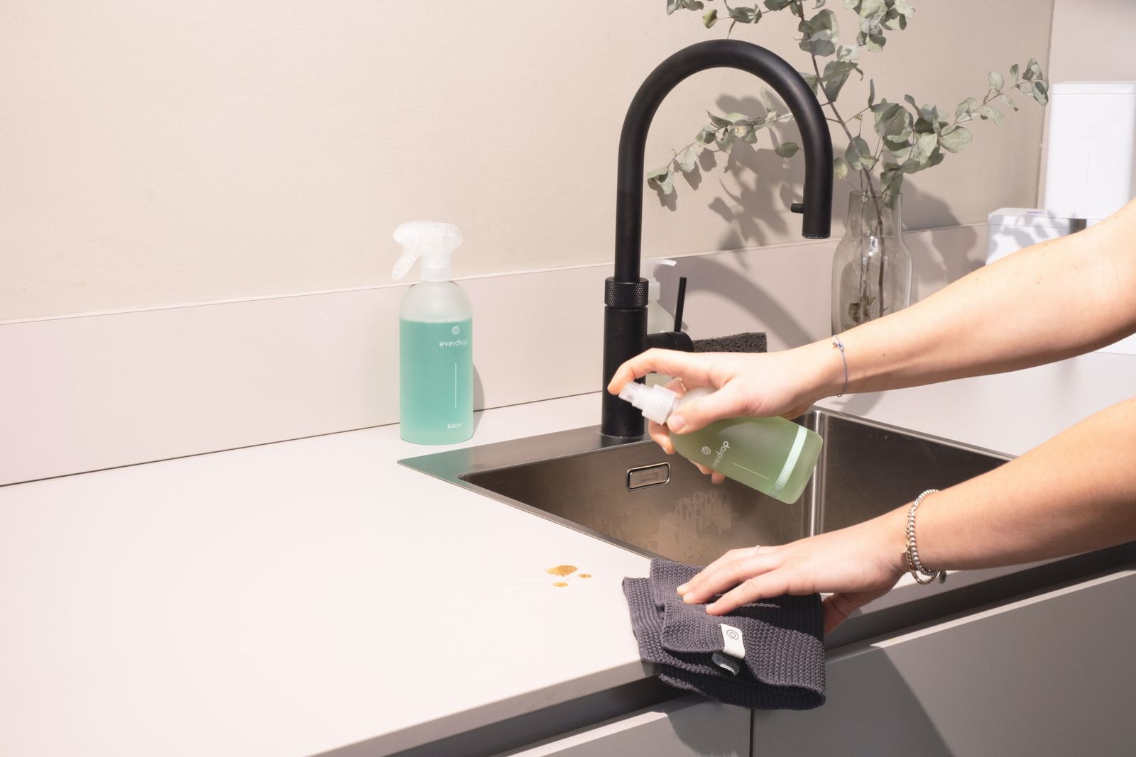 Hands wiping a black kitchen sink and chrome faucet with a microfiber cloth and cleaning spray.