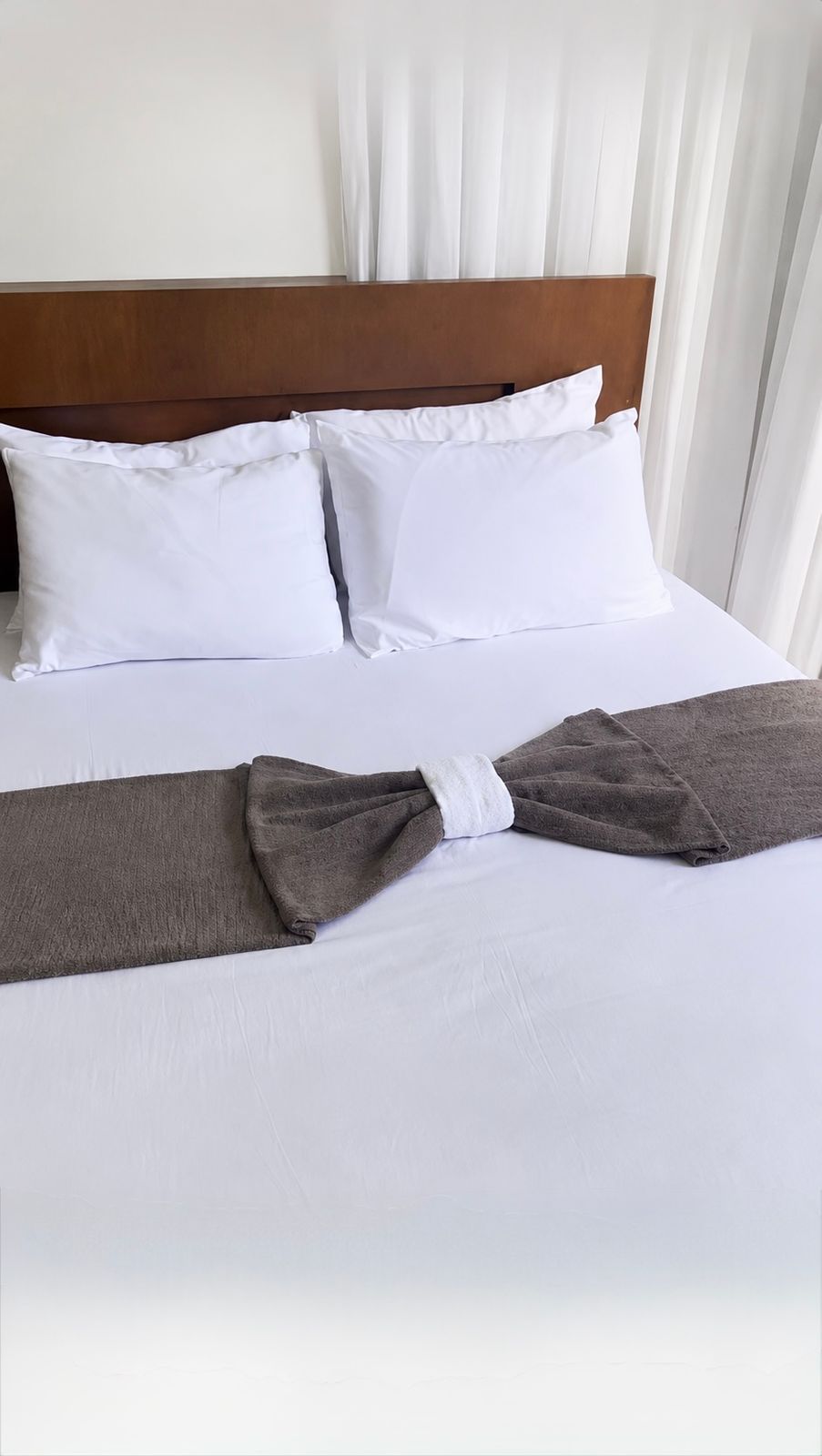 A neatly made bed with white pillows and sheets, a wooden headboard and a grey throw beside bright white curtains.