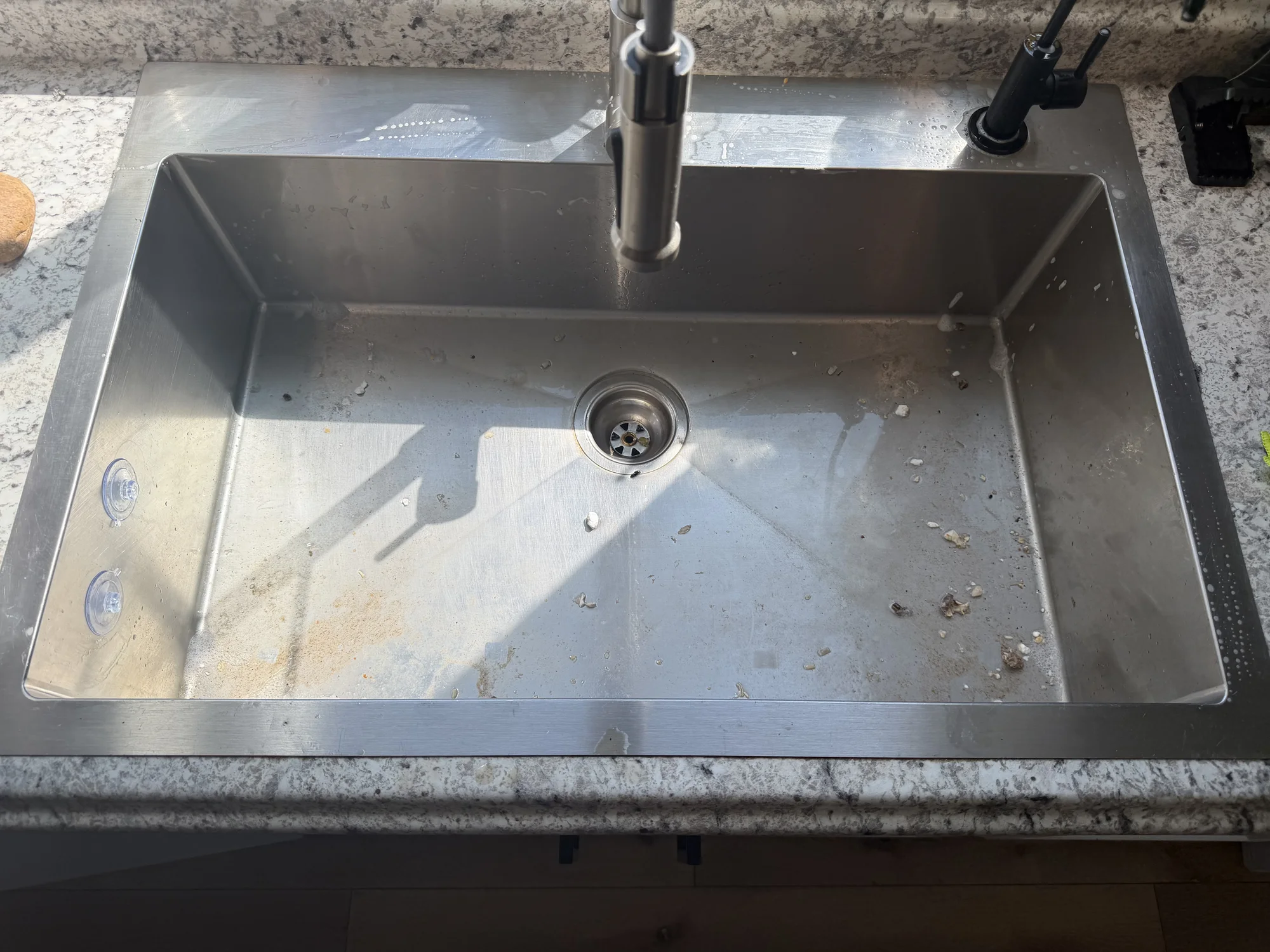A stainless steel undermount kitchen sink with food debris, water spots and residue in the basin.